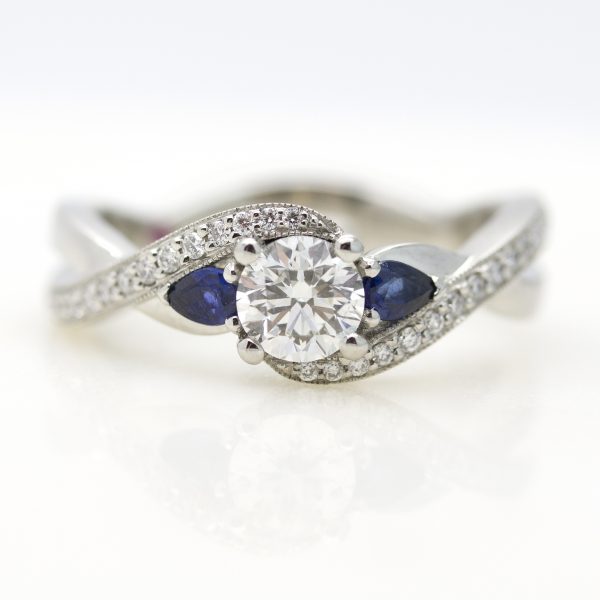 round diamond with diamond pave sweeping band and accent pear blue sapphires engagement ring
